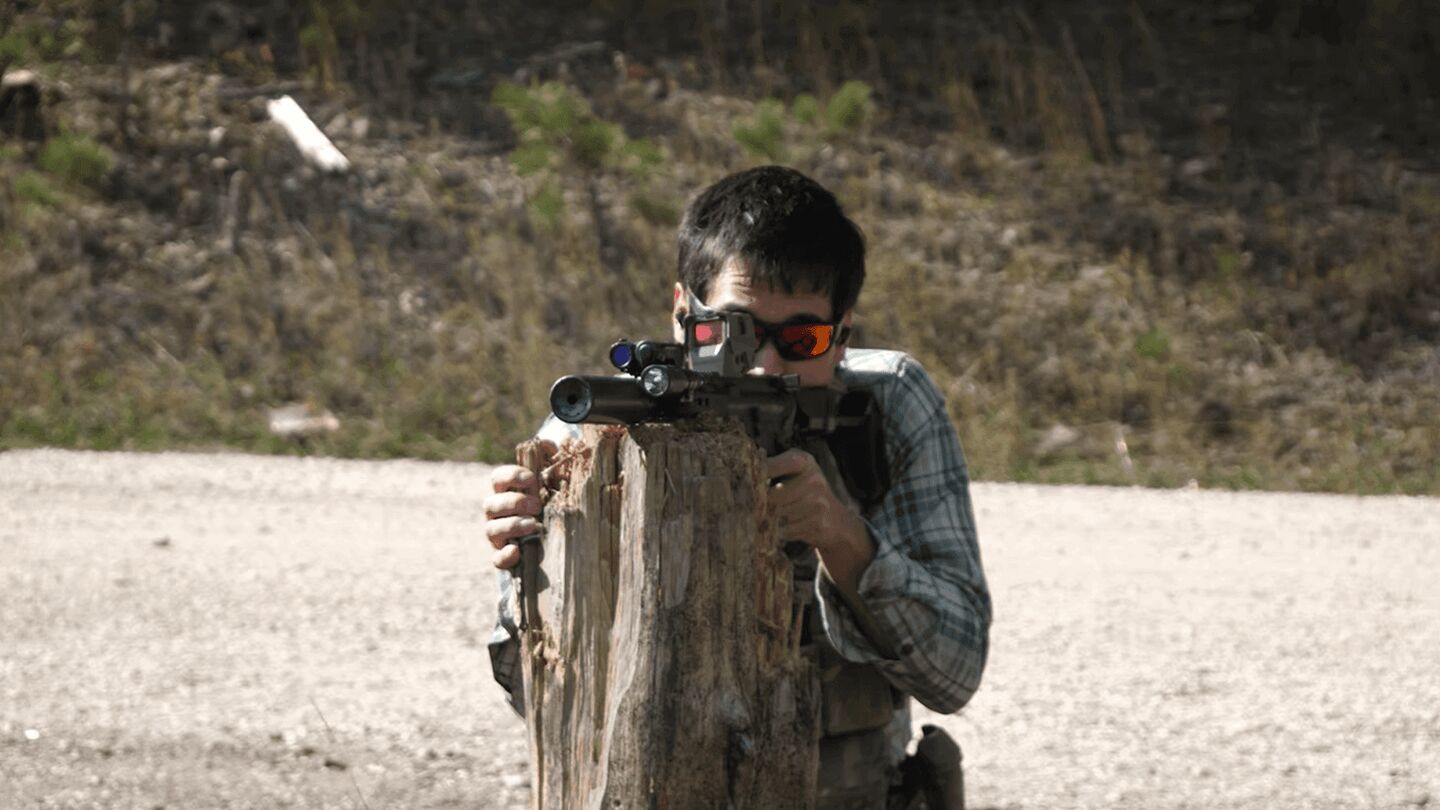 Shooter aims suppressed rifle over stump